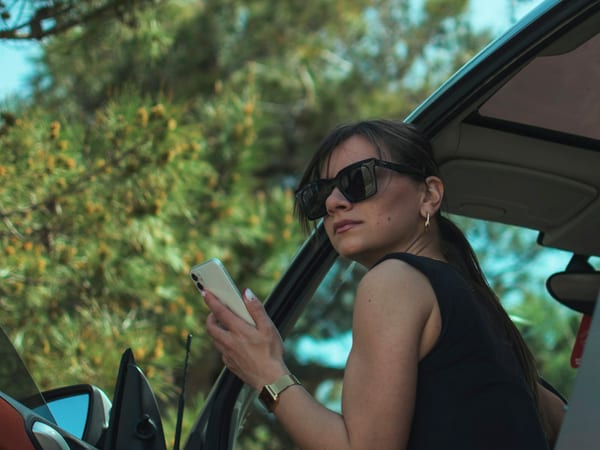 A woman in sunglasses at the open door of her car, phone in hand.