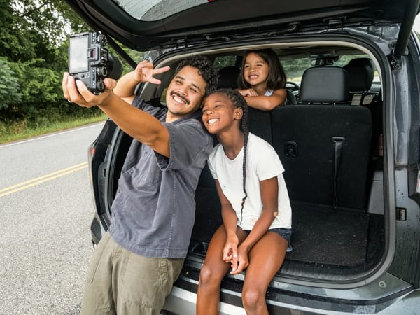 A father taking a selfie with his two daughters at the open boot of their car.